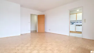 Leeres Wohnzimmer mit hellem Laminatboden und Zugang zu einer Küche.