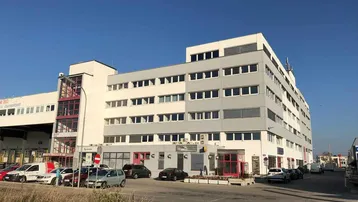 Modernes Bürogebäude mit mehreren Etagen und Parkplätzen davor unter blauem Himmel.