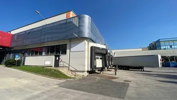Außenansicht eines Industriegebäudes mit Laderampen und einem Lastwagen auf dem asphaltierten Hof unter blauem Himmel.