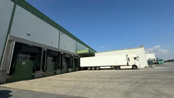 Außenansicht eines großen Lagergebäudes mit mehreren Laderampen und einem parkenden LKW unter blauem Himmel.