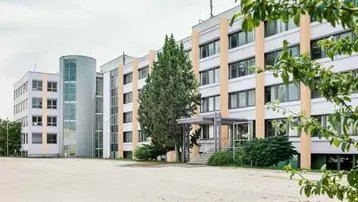 Modernes Bürogebäude mit mehreren Etagen, großen Fenstern und einem gläsernen Treppenhaus, umgeben von Bäumen und einem Parkplatz.