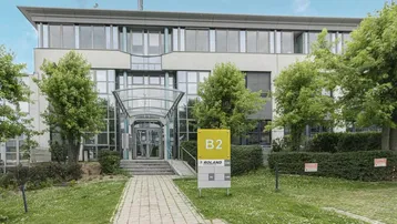 Modernes Bürogebäude mit Glasfassade, umgeben von Grünflächen und Bäumen, mit einem klaren Eingangsbereich.
