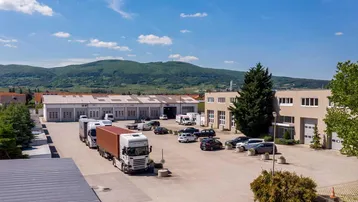 Luftaufnahme eines Gewerbegebiets mit mehreren Gebäuden, einem großen Parkplatz und Lastwagen vor einer Bergkulisse.