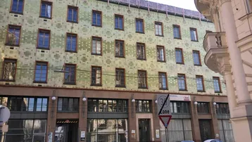 Historisches Gebäude mit markanter grüner Kachelfassade und Geschäften im Erdgeschoss an einer Stadtstraße.
