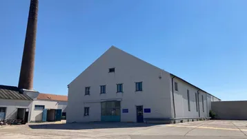 Großes Industriegebäude mit Wellblechfassade und hohem Schornstein unter klarem blauem Himmel.