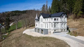 Historische Villa in Hanglage mit moderner Erweiterung und Wald im Hintergrund.