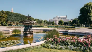 Idyllischer Park mit Springbrunnen, Teich und blühenden Blumenbeeten an einem sonnigen Tag.
