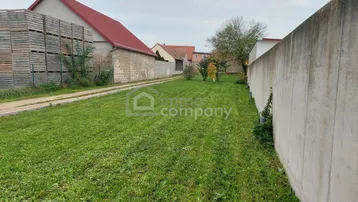 Grünfläche mit angrenzender Betonmauer und Gebäuden im ländlichen Hintergrund.