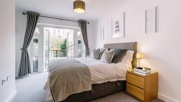 Helles Schlafzimmer mit Doppelbett, Teppichboden und Zugang zu einem französischen Balkon mit Blick ins Grüne.