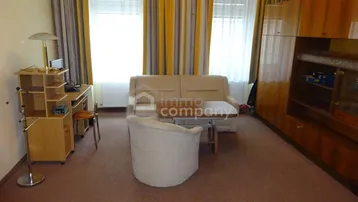 Geräumiges Wohnzimmer mit Teppichboden, Sofa, Schreibtisch und großen Fenstern mit Vorhängen.