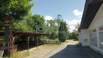 Außenbereich mit gepflastertem Weg, einem überdachten Unterstand und einem älteren Gebäude unter blauem Himmel.