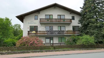Weißes Mehrfamilienhaus mit mehreren Balkonen und traditionellen Holzgeländern, umgeben von Bäumen und Sträuchern.
