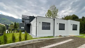 Modernes Einfamilienhaus mit heller Fassade, dunklen Fensterläden und gepflegtem Vorgarten vor Bergkulisse.
