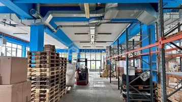 Geräumige Lagerhalle mit hohen Decken, blauen Stützbalken, Lüftungsrohren und Palettenregalen.