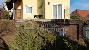Gelbes Einfamilienhaus mit braunem Holzdach und kleiner Terrasse im Vorgartenbereich.