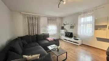 Geräumiges Wohnzimmer mit dunklem Ecksofa, Holzboden und zwei Fenstern, die für natürliches Licht sorgen.