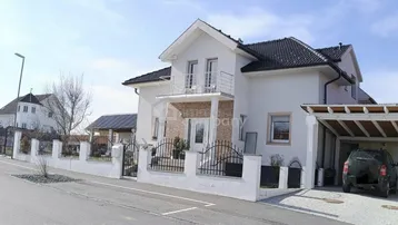 Weißes Einfamilienhaus mit dunklem Dach, Balkon und Carport an einer ruhigen Straße.
