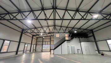 Große, helle Industriehalle mit hoher Decke, freiliegenden Stahlträgern und einem Zwischengeschoss mit Treppenzugang.