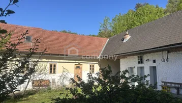 Zwei traditionelle Gebäude mit roten Ziegeldächern und viel Grün in ländlicher Umgebung unter blauem Himmel.