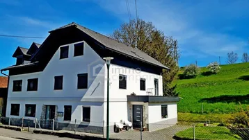 Weißes Wohnhaus mit dunklem Dach und grüner Hügellandschaft im Hintergrund unter blauem Himmel.