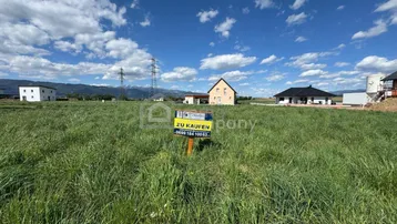 Weites grünes Feld mit einem 'Zu Kaufen'-Schild, umgeben von Häusern und Bergen im Hintergrund.
