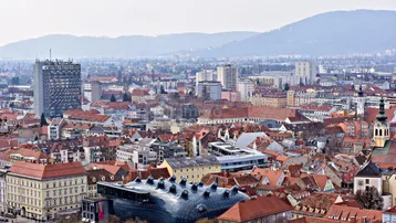 Panoramablick über eine Stadt mit roten Dächern und Bergen im Hintergrund, inklusive moderner Architektur.