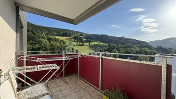 Heller Balkon mit Fliesenboden, rotem Geländer und weitem Blick ins Grüne.
