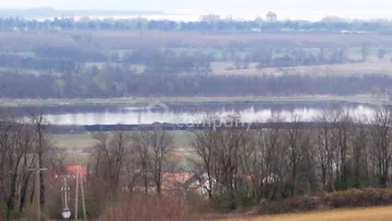 Weitläufige Landschaft mit Feldern, Bäumen und einem See im Hintergrund, unter einem bewölkten Himmel.