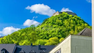 Blick auf eine grüne Hügellandschaft mit einer Burgruine im Hintergrund und Gebäuden im Vordergrund unter blauem Himmel.