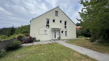 Modernes Einfamilienhaus mit heller Fassade und gepflegtem Vorgarten unter bewölktem Himmel.