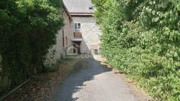 Ein schmaler Schotterweg führt zu einem alten Steingebäude, umgeben von dichtem Grün.