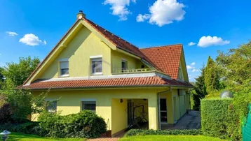 Gelbes Einfamilienhaus mit rotem Ziegeldach, umgeben von einem gepflegten Garten unter blauem Himmel.