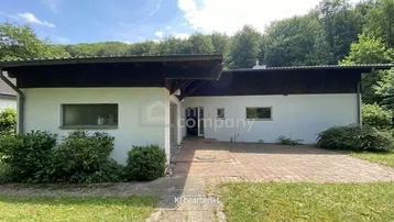 Einfamilienhaus mit weißer Fassade, dunklem Flachdach und gepflastertem Eingangsbereich, umgeben von viel Grün.