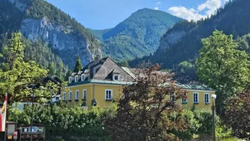 Gelbes Gebäude mit Mansarddach, umgeben von grünen Bäumen und majestätischen Bergen unter blauem Himmel.
