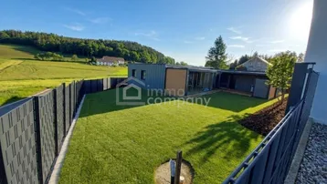 Moderner Neubau mit gepflegtem Rasen und Blick auf die umliegende Landschaft und Hügel.
