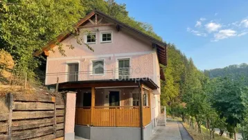 Hausfassade mit Balkonen und Holzelementen, eingebettet in eine grüne Hügellandschaft unter blauem Himmel.