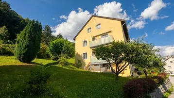 Gelbes Mehrfamilienhaus mit grüner Umgebung und blauer Himmel unter strahlender Sonne.