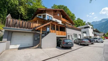 Modernes Mehrfamilienhaus mit Holzverkleidung, Garagen und Parkplätzen vor einer bewaldeten Bergkulisse.