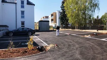 Außenansicht eines modernen Neubaus mit einem großen, asphaltierten Parkplatz und Bäumen.