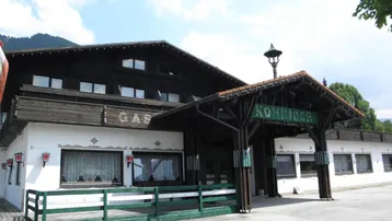 Traditionelles Gebäude mit dunkler Holzfassade und weißem Putz, umgeben von Bergen und Natur.
