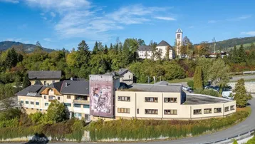 Luftaufnahme eines Gebäudekomplexes in ländlicher Umgebung mit Kirche im Hintergrund und bewaldeten Hügeln.