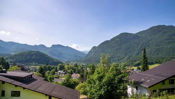 Panoramablick auf eine grüne Berglandschaft mit Häusern in einem Tal unter blauem Himmel.