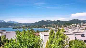 Weitläufiger Ausblick auf den See, die umliegenden Berge und die Stadt mit modernen Gebäuden im Vordergrund.