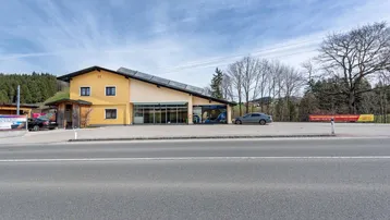 Gebäudeansicht mit großem Parkplatz, Photovoltaikanlage auf dem Dach und umliegender Landschaft.