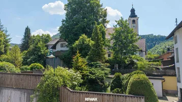 Malerische Umgebung mit einem Kirchturm, üppigem Grün und traditionellen Häusern unter blauem Himmel.