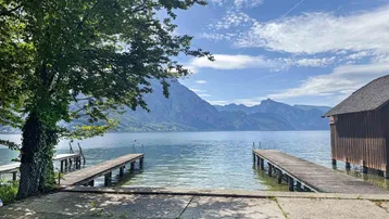 Idyllischer Seeblick mit Bergen im Hintergrund, umgeben von grüner Natur und Holzstegen.