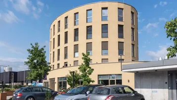 Modernes, rundes Gebäude mit beiger Fassade und Parkplätzen davor unter blauem Himmel.