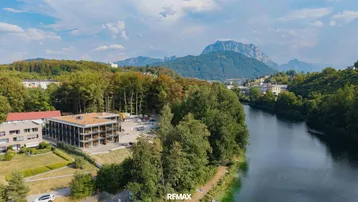 Luftaufnahme eines modernen Gebäudes am Flussufer mit üppiger Vegetation und majestätischem Bergblick.