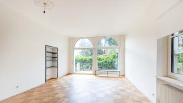 Helles Wohnzimmer mit Fischgrätparkett, großen Bogenfenstern und Zugang zu einem Balkon mit Grünblick.