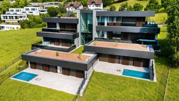 Moderne Architektur mit mehreren Ebenen, Flachdächern, großen Fenstern und kleinen Pools, umgeben von Grünflächen.